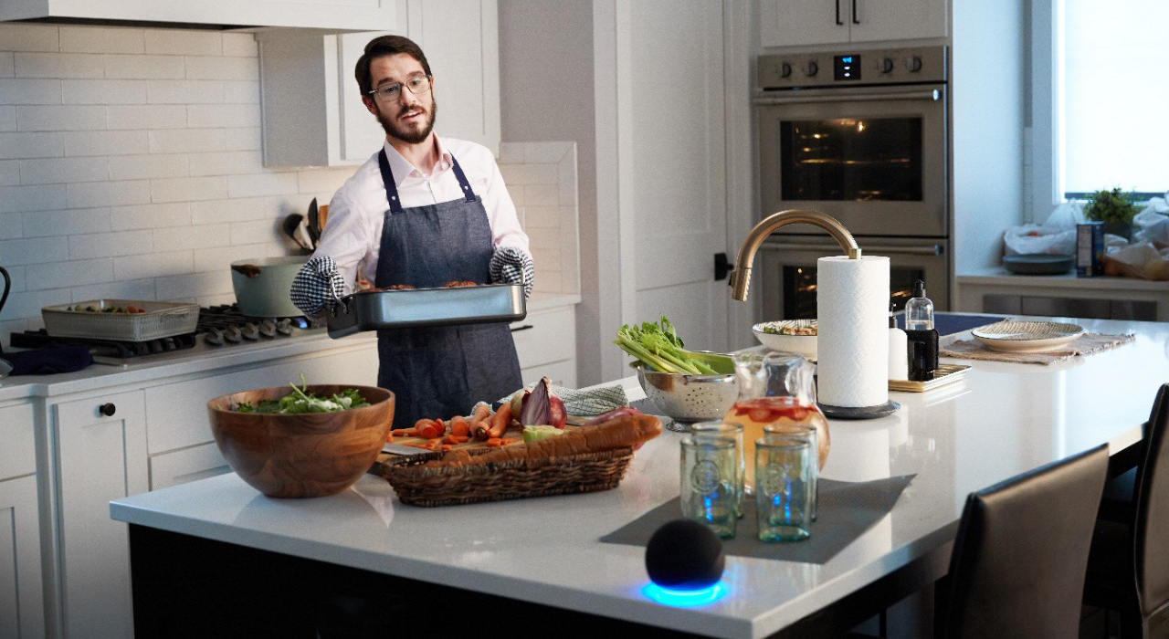 Man holding a baking pan in a busy home kitchen as he uses an Alexa voice command to unlock his smart lock.