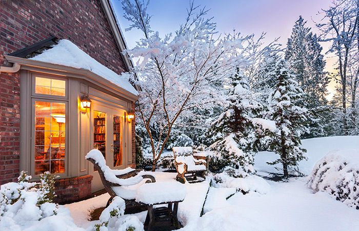 Snowy scene featuring a home exterior lit up from inside. Outside patio furniture is covered in snow.