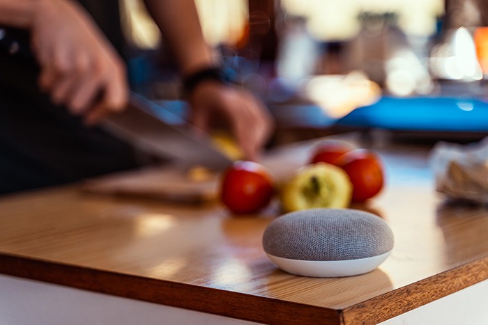 Smart speaker on table while peson in background is chopping vegetables.