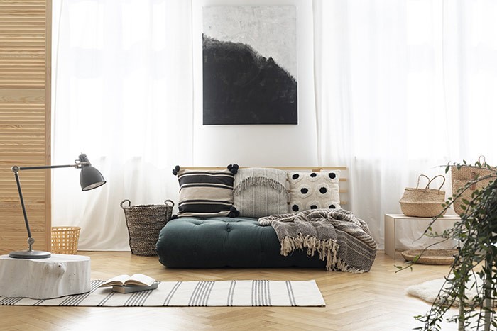 Poster above green futon with patterned pillows in bright living room interior with lamp. Real photo